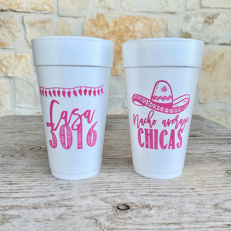 May include: Two white foam cups featuring pink printed designs. The left cup displays a string of lights above the text "fasa 3016". The right cup features a sombrero graphic above the text "Nacho average CHICAS" in a mix of script and block fonts.