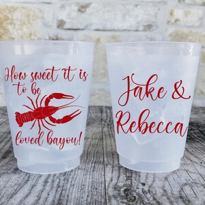 May include: Two clear plastic cups with ice. One cup has the text "How sweet it is to be loved bayou!" and a red crawfish graphic. The other cup says "Jake & Rebecca" in red script.
