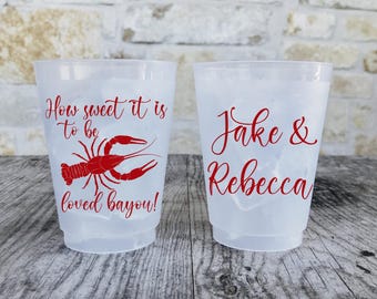 May include: Two clear plastic cups with ice. One cup has the text "How sweet it is to be loved bayou!" and a red crawfish graphic. The other cup says "Jake & Rebecca" in red script.