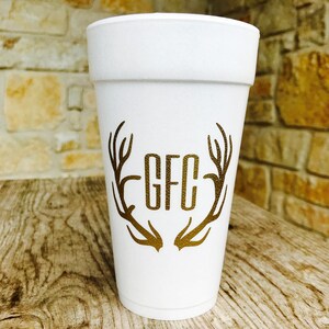 May include: A white foam cup with a gold antler design and the letters "GFC". The cup is sitting on a wooden surface, with a brick wall in the background. The cup is a standard size.