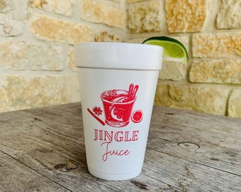 Puede incluir: Un vaso de espuma blanco con un gráfico rojo de una copa de cóctel con ramas de canela y una rodaja de cítrico. El texto JINGLE Juice está impreso en rojo debajo de la ilustración. Un gajo de lima verde fresco descansa sobre el borde del vaso.