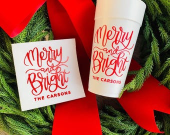 May include: A white cup and a white square card with the words "Merry and Bright" in red script, along with "The Carsons." The cup and card are set against a green wreath with a large red bow.