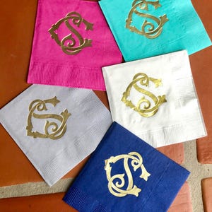 May include: A collection of personalized paper napkins in various colors, including pink, teal, white, gray, and blue. Each napkin features a gold monogram design. Ideal for events and celebrations.