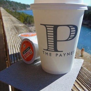 May include: A white paper coffee cup with a lid, featuring a large gray letter "P" and the words "THE PAYNES". Two coffee pods are next to the cup. The cup sits on a gray placemat, with a scenic background.
