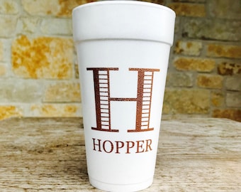 May include: A white foam cup with the name "HOPPER" printed in a copper-colored font. The letter "H" is prominently displayed above the name. The cup is sitting on a wooden surface.