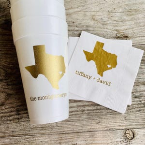May include: White foam cups and napkins with gold foil Texas state designs. The cups are printed with "the montgomerys" and the napkins with "tiffany + david". The design includes a heart shape within the Texas outline.
