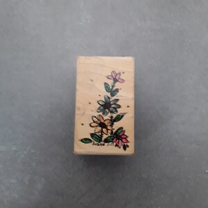 May include: A rectangular wooden stamp featuring a floral design with pink, blue, and orange flowers. The stamp has a handwritten signature that reads "SUSAN JILL". The flowers are arranged on a stem with green leaves.