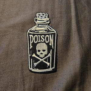 Pode incluir: Etiqueta bordada em preto e branco de uma garrafa de veneno com uma caveira e ossos cruzados. A garrafa está etiquetada como "POISON".