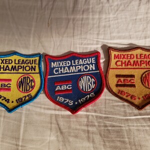 May include: Three embroidered patches with a shield shape. Each patch features the text "MIXED LEAGUE CHAMPION", the letters "ABC" and "WIBE", and the years 1974-1975, 1975-1976, and 1976-1977. The patches have blue, red, and brown borders.