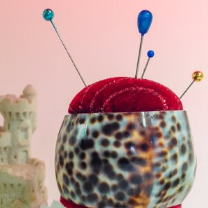 May include: A pincushion made of a brown and white shell with a red velvet top. The pincushion is sitting on a clear glass base and has several colorful pins sticking out of it. There is a small sandcastle in the background.