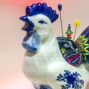 May include: A white ceramic rooster figurine with blue and white painted details. The rooster has a blue crest and a black, blue, and yellow fabric tail with a red star on top.