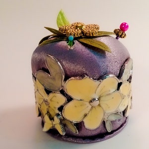 May include: A purple velvet pincushion with yellow flowers and green leaves. The pincushion has a gold and beaded embellishment on top.