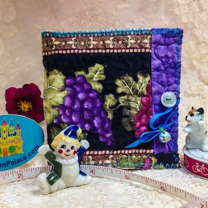 May include: A small quilted fabric pouch with a grapevine design and a blue ribbon. The pouch has a button closure and is decorated with a small blue button and a bead. The pouch is sitting on a white surface with a measuring tape, a clown figurine, and a sticker with the text "PincushionPalace.com".