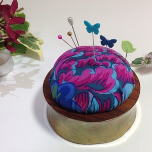 May include: A round pincushion with a floral print in shades of pink, blue, and green. The pincushion is on a wooden base with a brass finish. There are several colorful butterfly pins stuck in the pincushion.
