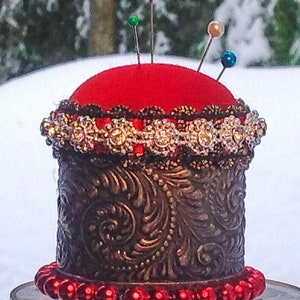 May include: A red velvet pincushion with a gold metal base and a decorative beaded trim. The pincushion is sitting on a glass pedestal.