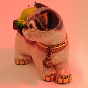 May include: White ceramic cat figurine with a black and blue design. The cat is wearing a gold and pink collar with a tassel. The cat has a yellow and green pincushion on its back with colorful pins.
