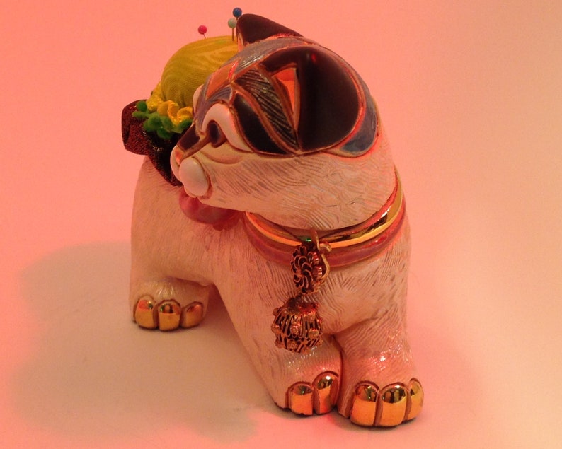 May include: White ceramic cat figurine with a black and blue design. The cat is wearing a gold and pink collar with a tassel. The cat has a yellow and green pincushion on its back with colorful pins.