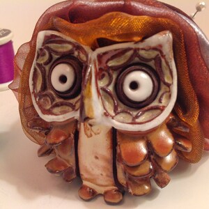 May include: A ceramic owl pincushion with brown and white details, wearing a brown and orange ribbon. The owl has large, white eyes with black pupils.