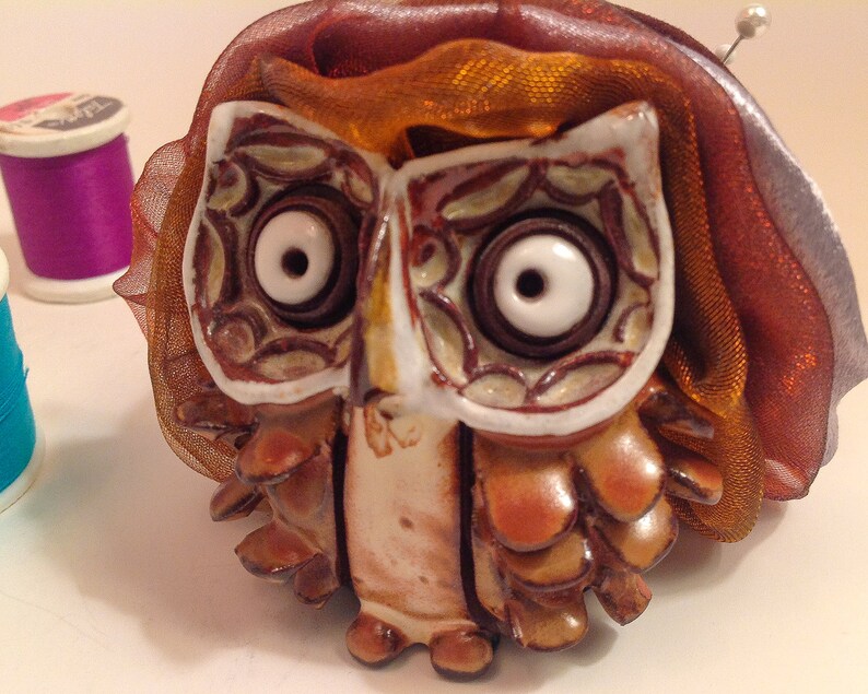 May include: A ceramic owl pincushion with brown and white details, wearing a brown and orange ribbon. The owl has large, white eyes with black pupils.