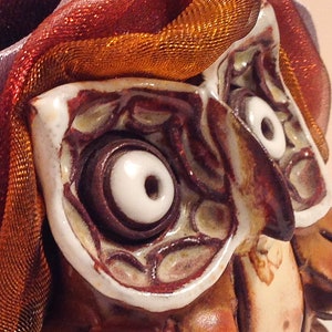May include: Close-up of a ceramic owl sculpture with white eyes and brown details. The owl is decorated with a red and orange fabric.