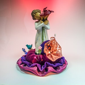 May include: A white figurine of a person holding a bouquet of purple and orange flowers, standing on a purple and orange pincushion with a blue bird on the side.