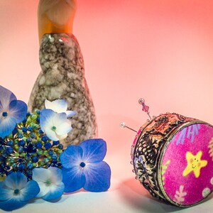 Op de afbeelding: Een decoratief speldenkussen met een roze stoffen bovenkant met een gele lachende ster en een zilveren filigraan basis. Blauwe en witte hortensia bloemen en een gespikkelde keramische vaas staan ook in de afbeelding.