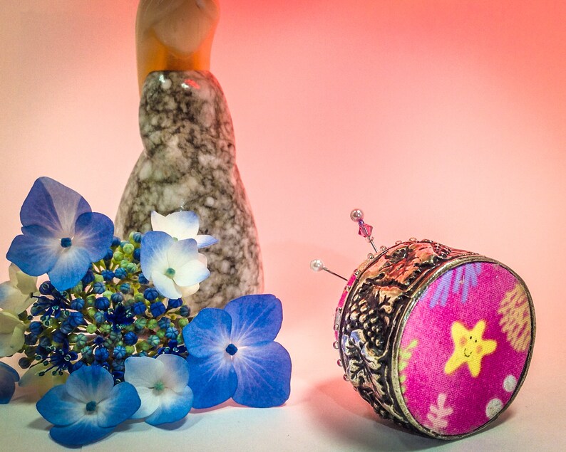 Op de afbeelding: Een decoratief speldenkussen met een roze stoffen bovenkant met een gele lachende ster en een zilveren filigraan basis. Blauwe en witte hortensia bloemen en een gespikkelde keramische vaas staan ook in de afbeelding.
