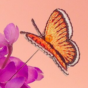 May include: A small orange and black embroidered butterfly with a white border. The butterfly is attached to a pin and is sitting on a purple flower.