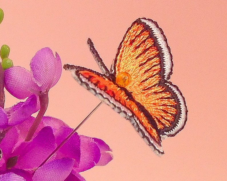 May include: A small orange and black embroidered butterfly with a white border. The butterfly is attached to a pin and is sitting on a purple flower.