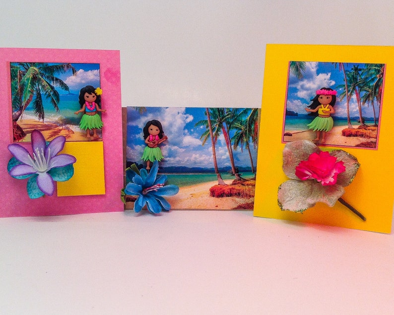 May include: Three handmade greeting cards with tropical beach scenes and hula girl figurines. Each card features a different colored background, a flower embellishment, and a tropical scene with palm trees and a beach.