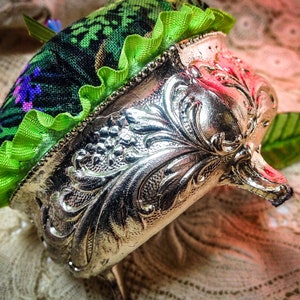 May include: A vintage silver pincushion with an ornate design and a green and purple fabric top with a ruffled trim.
