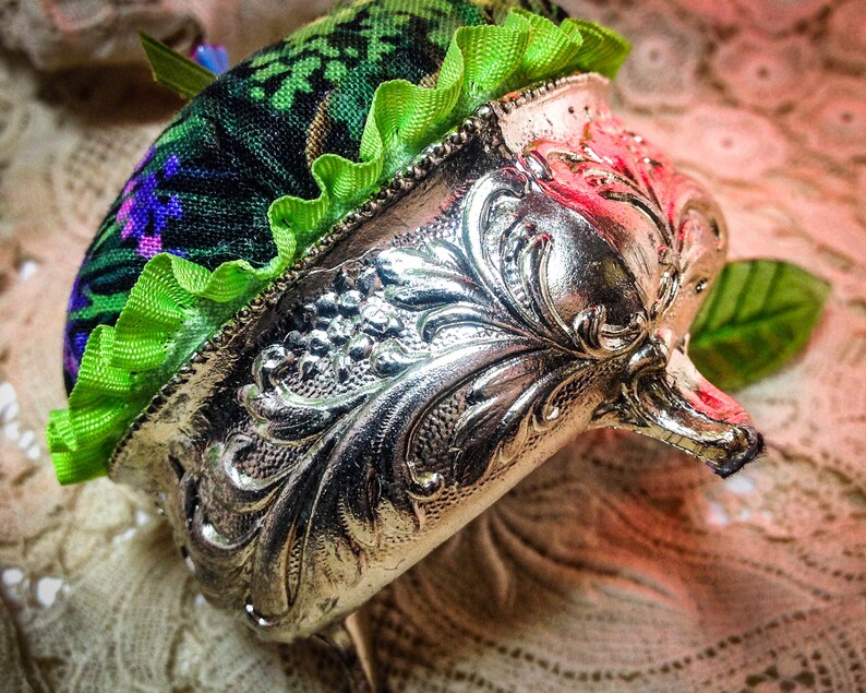 May include: A vintage silver pincushion with an ornate design and a green and purple fabric top with a ruffled trim.