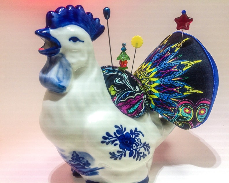 May include: A white ceramic rooster figurine with blue and white painted details. The rooster has a blue crest and a black, blue, and yellow fabric tail with a red star on top.