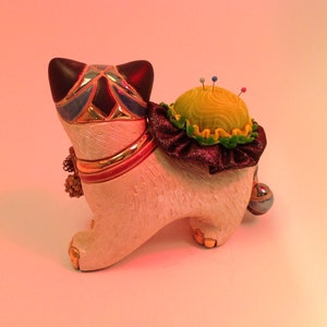 May include: A white ceramic cat figurine with a colorful mosaic design on its head and a yellow pincushion on its back. The pincushion has a green and purple ruffle around the bottom.