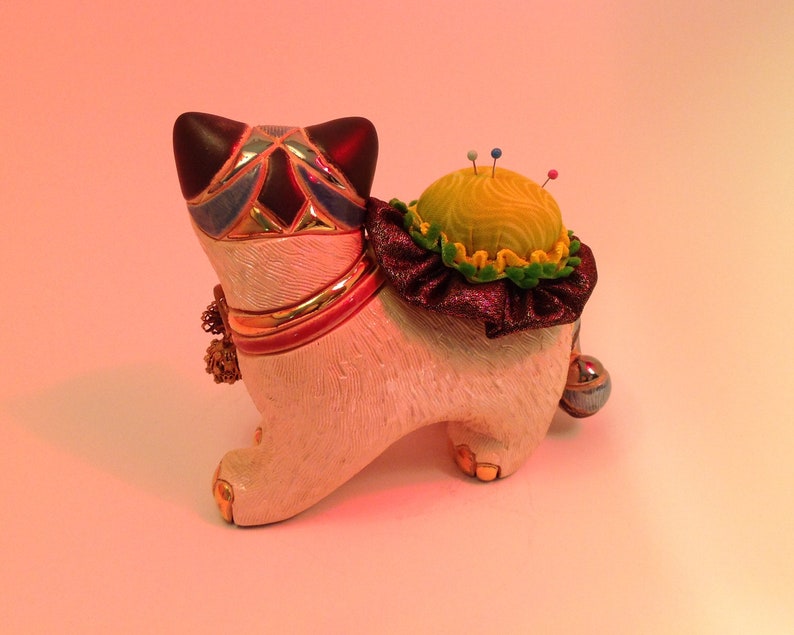 May include: A white ceramic cat figurine with a colorful mosaic design on its head and a yellow pincushion on its back. The pincushion has a green and purple ruffle around the bottom.