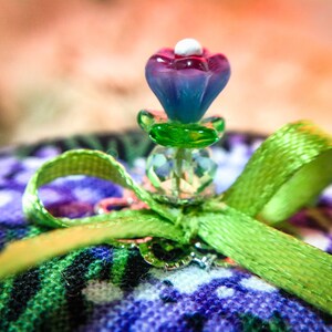 May include: A green satin ribbon tied into a bow with a purple and pink glass flower embellishment on top. The flower has a white center and is surrounded by clear glass beads.