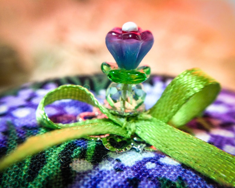 May include: A green satin ribbon tied into a bow with a purple and pink glass flower embellishment on top. The flower has a white center and is surrounded by clear glass beads.