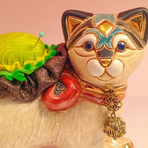 May include: A ceramic cat figurine with a green pincushion on its back. The cat has a white body with black and blue markings, gold accents, and blue eyes. It is wearing a red and gold collar with a gold charm.