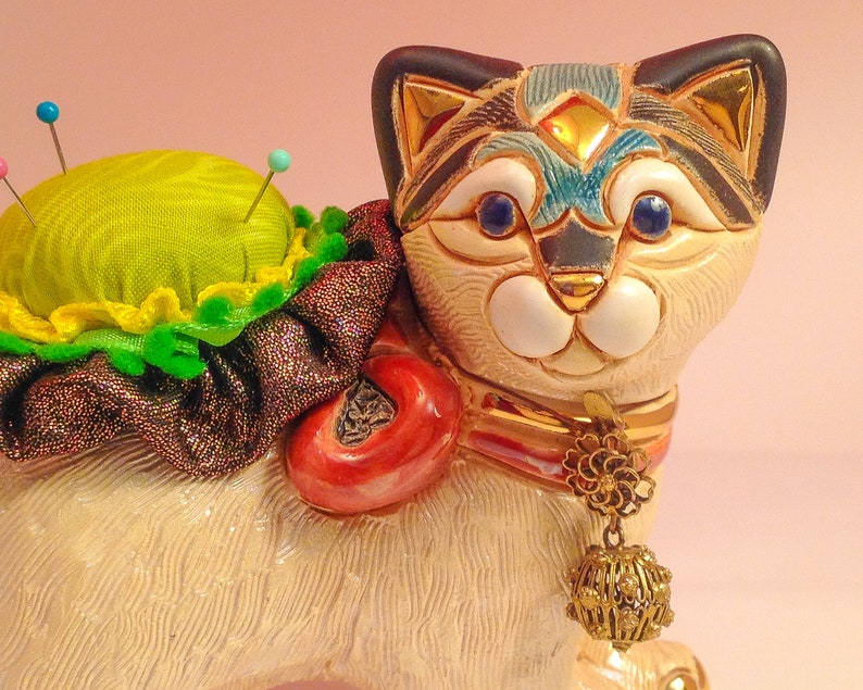 May include: A ceramic cat figurine with a green pincushion on its back. The cat has a white body with black and blue markings, gold accents, and blue eyes. It is wearing a red and gold collar with a gold charm.