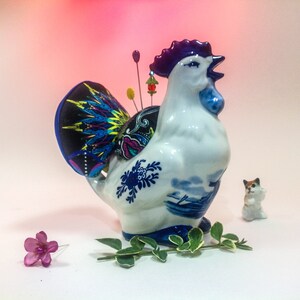 May include: A white ceramic rooster figurine with blue and white Delftware designs. The rooster has a purple comb and wattle and is holding a pincushion with colourful fabric and pins. A small white cat figurine is in the background.