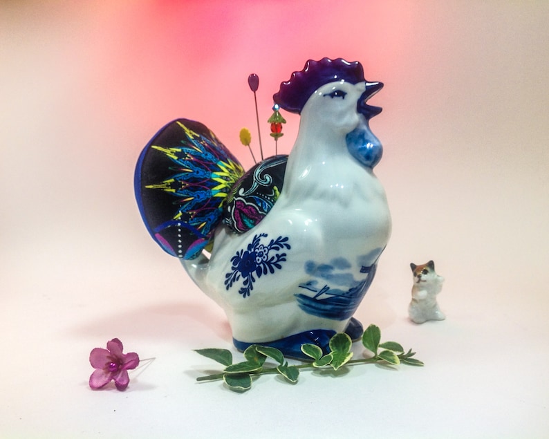 May include: A white ceramic rooster figurine with blue and white Delftware designs. The rooster has a purple comb and wattle and is holding a pincushion with colourful fabric and pins. A small white cat figurine is in the background.