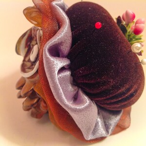 May include: A brown velvet pincushion with a purple satin ribbon and a floral embellishment. The pincushion is decorated with a small red pin and white pearl pins.