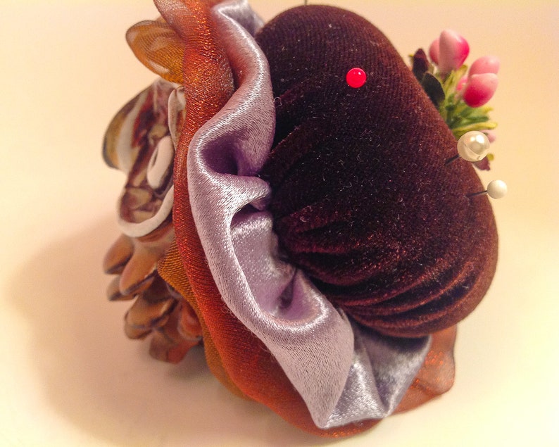 May include: A brown velvet pincushion with a purple satin ribbon and a floral embellishment. The pincushion is decorated with a small red pin and white pearl pins.