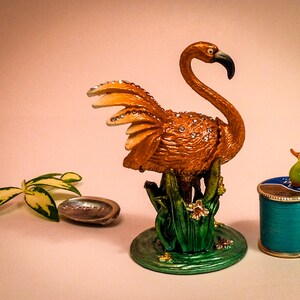 May include: A gold-colored flamingo figurine with rhinestones, standing on a green base with flowers. A small green duck figurine sits on top of a teal blue spool of thread.