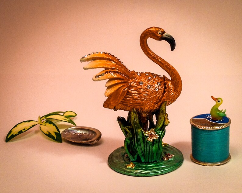 May include: A gold-colored flamingo figurine with rhinestones, standing on a green base with flowers. A small green duck figurine sits on top of a teal blue spool of thread.