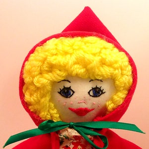 May include: A handmade doll with a red hooded cloak, yellow yarn hair, and a white face with embroidered features. The doll is wearing a green ribbon around its neck.
