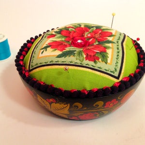 May include: A green and red floral fabric pincushion with black trim sits on top of a black and gold painted wooden bowl. The bowl has a floral design.