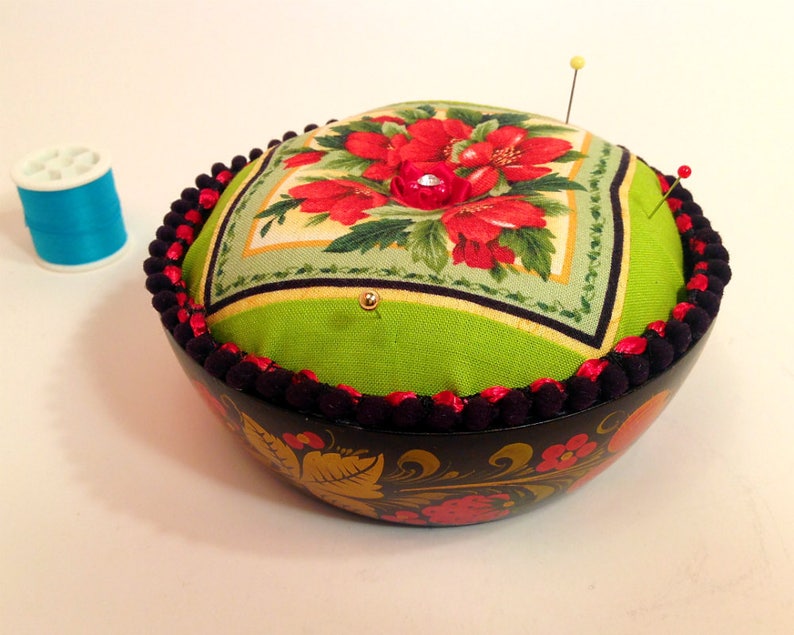 May include: A green and red floral fabric pincushion with black trim sits on top of a black and gold painted wooden bowl. The bowl has a floral design.