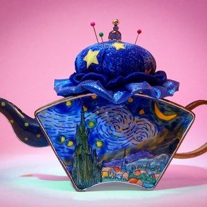 May include: A blue and white teapot with a pincushion lid featuring a starry night design. The pincushion is blue with a yellow star and gold trim. The teapot is decorated with a painting of a starry night sky with a crescent moon.