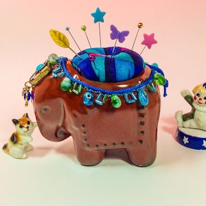 May include: A brown ceramic elephant pincushion with a blue and purple fabric pincushion on top. The elephant is decorated with colourful beads and a gold chain. A small porcelain clown figurine is on a blue and white base to the right of the elephant.